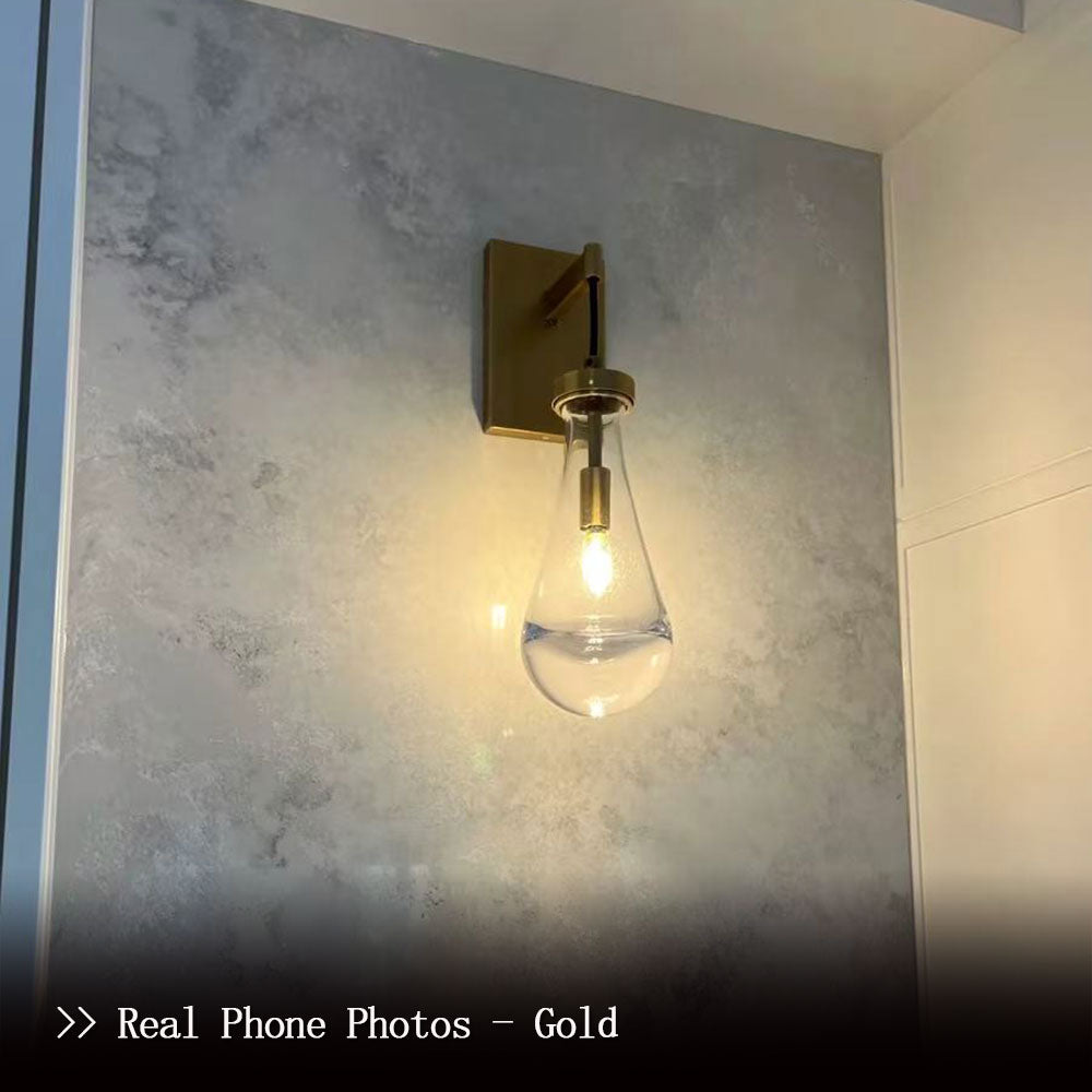 Customer real‑use photo of brass teardrop clear glass wall sconce, lit single wall lamp actual installation effect, brushed brass fixture with teardrop glass shade on textured interior wall