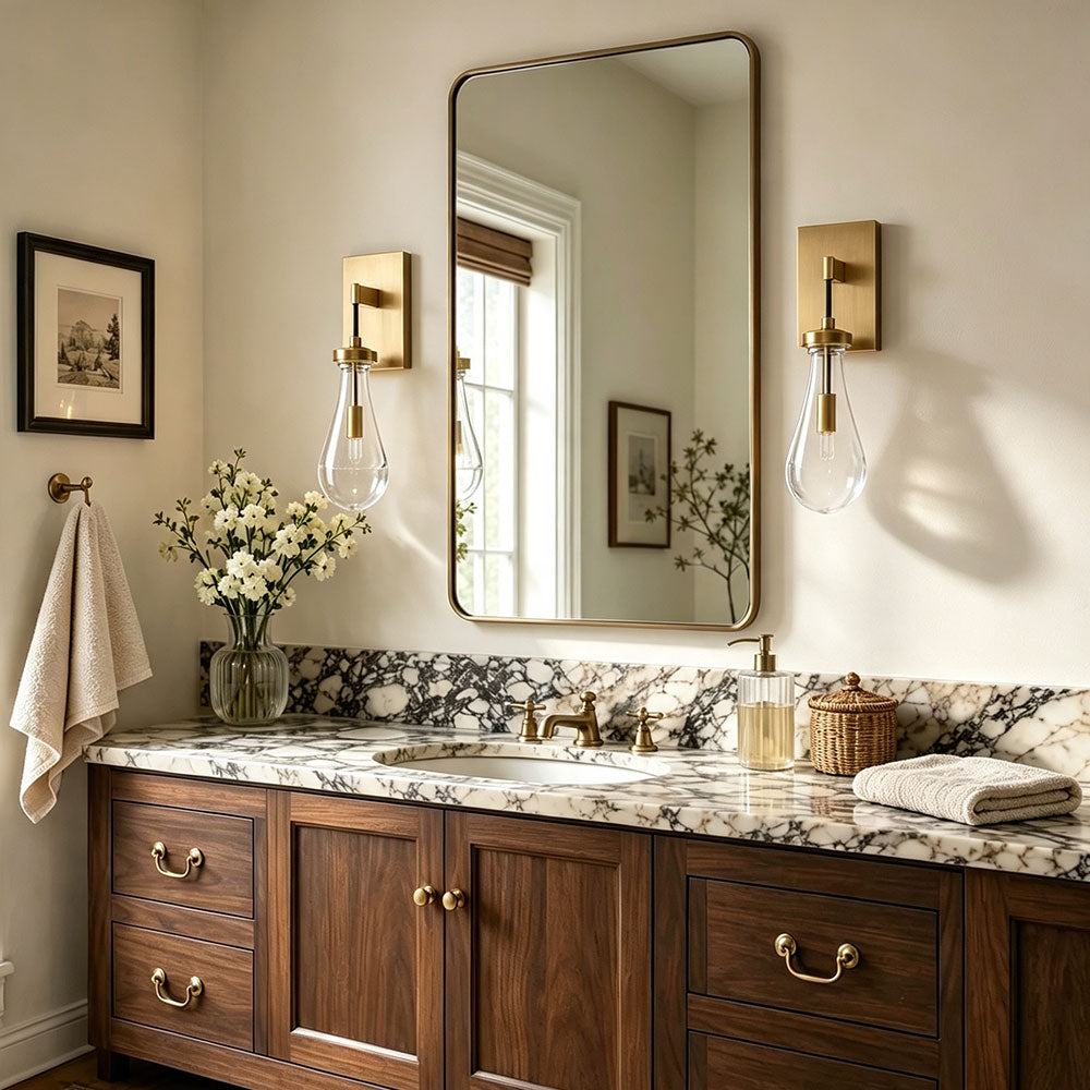Pair brushed brass teardrop clear glass wall sconces beside bathroom vanity mirror, modern farmhouse wall lamps for powder room, wooden cabinet marble countertop bathroom interior scene