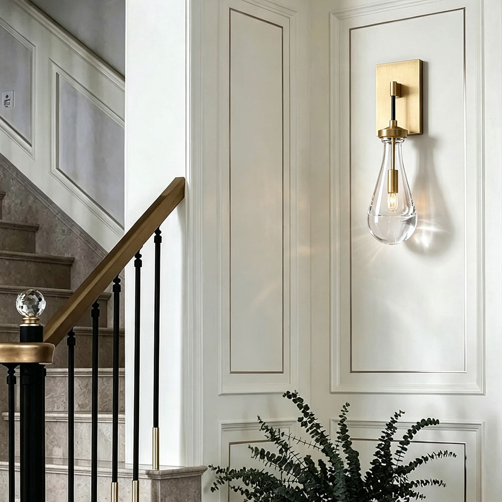 Single brushed brass teardrop clear glass wall sconce, lit wall light mounted on white panel wainscoting, modern vintage wall fixture for stairway hallway entry interior decor